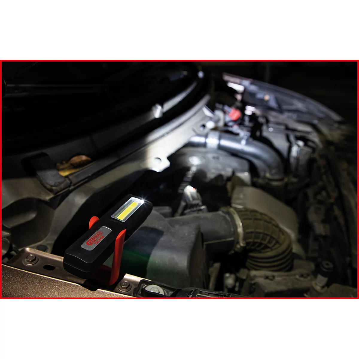 Een LED-werklamp verlicht de motorruimte van een auto. De lamp is bevestigd aan een rode clip.